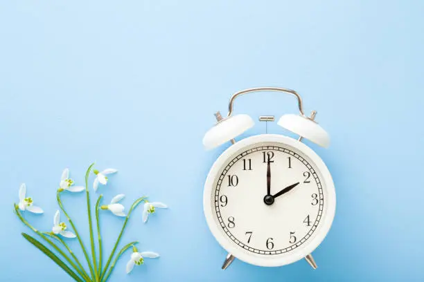 White alarm clock with fresh, beautiful snowdrops on light blue table background. First messengers of spring. Time concept. Closeup. Empty place for text. Top down view.