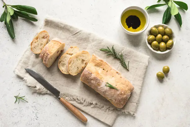 Ciabatta Bread with olive oil and green olives. Fresh Homemade Italian Bread Ciabatta slised on linen napkin, white table, top view.
