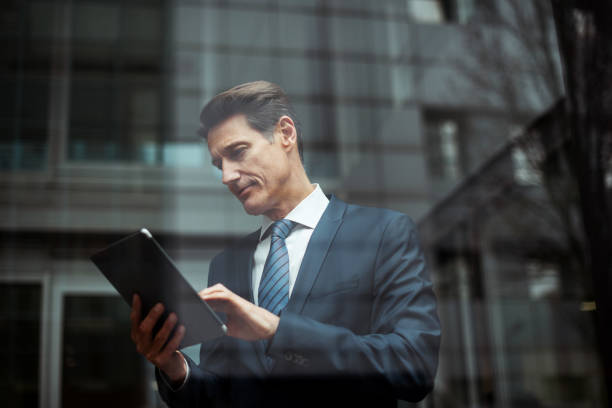 German businessman Formally dressed 55-year-old businessman is holding a digital tablet and looking through the window of the bright office. georgijevic frankfurt stock pictures, royalty-free photos & images