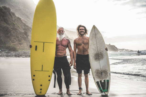 amigos multigeração indo para surfar na praia tropical - pessoas em família se divertindo fazendo esporte extremo - idosos alegres e conceito de estilo de vida saudável - surfing beach family father - fotografias e filmes do acervo