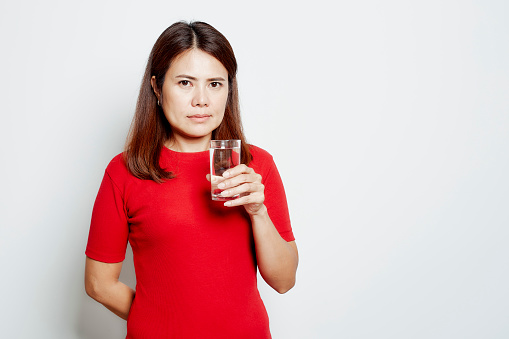 Young smiling asian woman drinks water from a glass isolated studio portrait on white background, Pure water concept and health care