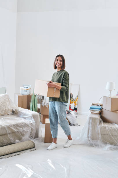 Young cheerful woman in casualwear carrying box while moving along room Young cheerful woman in casualwear carrying box with stuff while moving along living-room of new flat or house home ownership women stock pictures, royalty-free photos & images