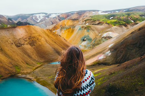 Young woman hiker with long hair and in a wool traditional Icelandic sweater got to the top of the mountain, relaxing looking at the beautiful multi-colored panoramic landscape with mountains and wild blue lakes in Landmannalaugar valley, Iceland