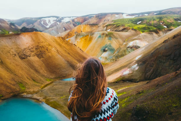 Woman traveler enjoying the view of scenic colorful rainbow mountains and turquoise lake in the wilderness Young woman hiker with long hair and in a wool traditional Icelandic sweater got to the top of the mountain, relaxing looking at the beautiful multi-colored panoramic landscape with mountains and wild blue lakes in Landmannalaugar valley, Iceland lava photos stock pictures, royalty-free photos & images