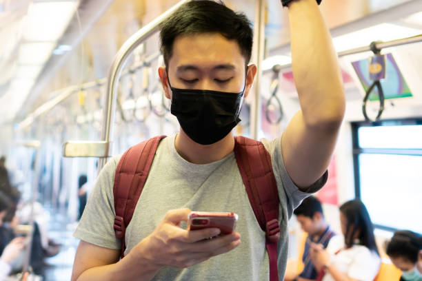 close up of one young asian man wearing a black surgical face mask and using a mobile phone in subway train during new type coronavirus covid-19 pneumonia outbreak and pm 2.5 smog air pollution crisis in big city - human face chinese ethnicity close up men imagens e fotografias de stock