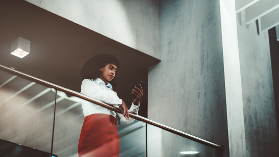 A pensive young African-American woman entrepreneur in a white shirt and red skirt is replying a message using her cellphone while leaning on chrome railing of a balcony in an office open-space area