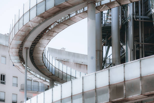 A close-up view of a contemporary construction of an oil refinery or a modern fuel factory facility in an industrial zone, with a two-section round bridge, multiple pipes, iron beams, and stairs