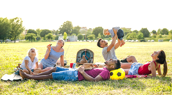 Young multiracial families having fun playing with kids at pic nic garden party - Multiethnic joy and love concept with mixed race people together with children at park barbecue - Warm bright filter