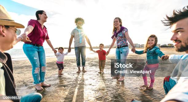 View point of young families dancing at beach on ring around the rosy style - Lifestyle joy concept with mixed race people having fun moment holding hands - Vivid backlight filter with sunshine halo