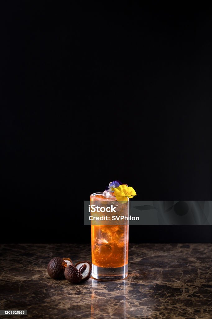 Cloudy Lemonade. The drink is located on a black background. Claudi lemonade drink stands on a granite slab. The drink is decorated with flowers. Berries of lychee are located next to the drink. Black Background Stock Photo