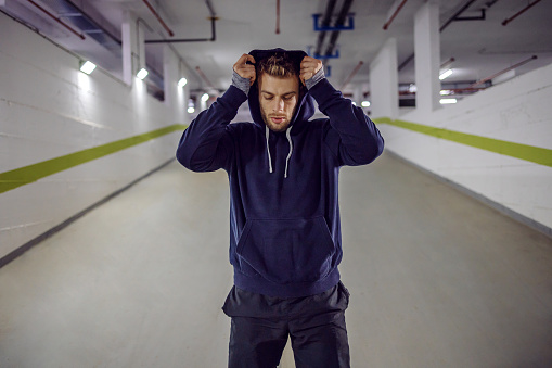 Young sportsman putting hoodie on head while standing in underground garage.