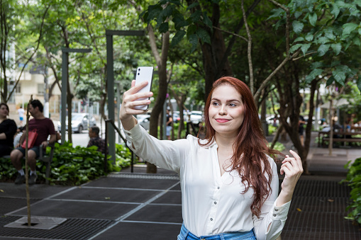 Happy beautiful redhead woman with white shirt making a selfie with a smartphone at the park, sunny day