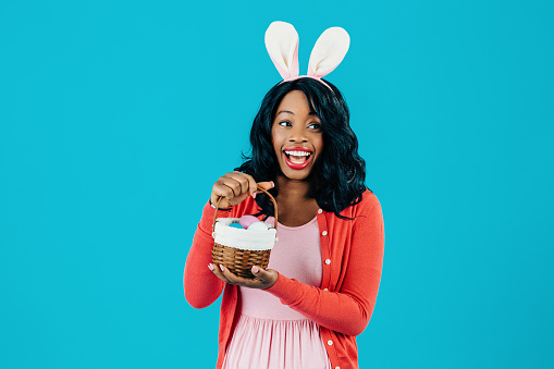 Portrait of an excited mother with Easter eggs basket and bunny ears looking to side, isolated on blue background