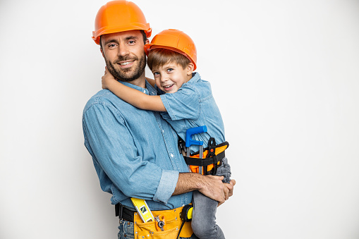 Smiling dad is holding boy on hands and hugging while they are wearing building helmets and belts with hand tools. Isolated on white background