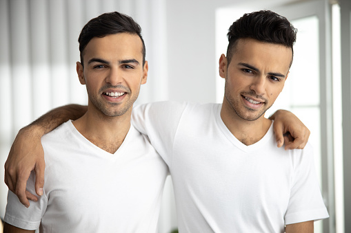 Waist up of happy friendly male twins in white t-shirts embracing at home stock photo