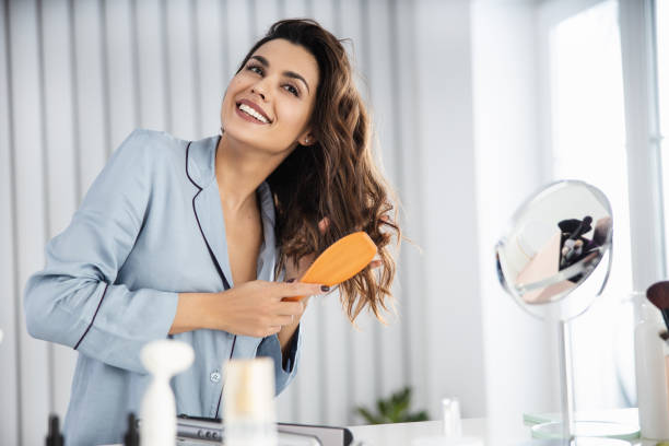 Beautiful young female brushing healthy hair at home Happy pretty woman in pajamas combing her hair n front of the mirror stock photo curling tongs photos stock pictures, royalty-free photos & images