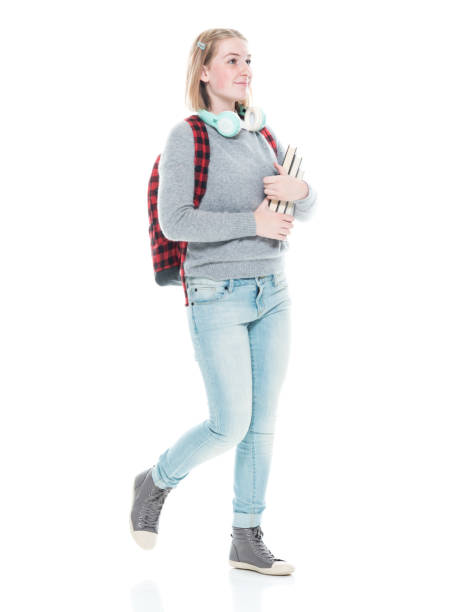 istock Generation z female university student walking in front of white background wearing backpack and holding book and using headphones 1209587141