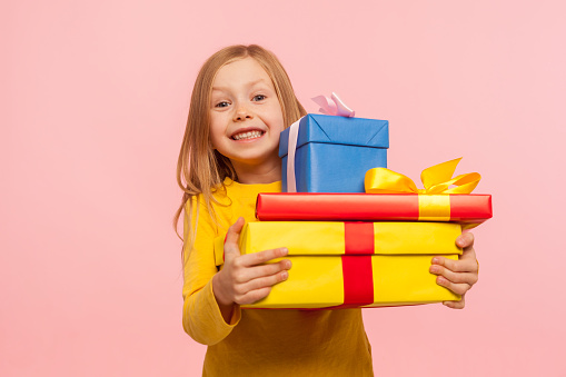 Delighted little girl embracing lot of gift boxes and smiling at camera with expression of sincere childish happiness, enjoying perfect birthday with many presents. indoor studio shot, pink background