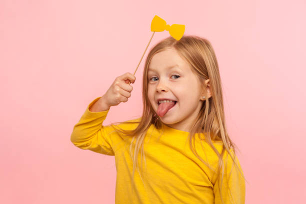 Portrait of playful adorable little ginger girl holding paper bow and sticking out tongue, naughty child having fun Portrait of playful adorable little ginger girl holding paper bow and sticking out tongue, naughty child having fun, wearing masquerade accessory. indoor studio shot isolated on pink background child tied up stock pictures, royalty-free photos & images
