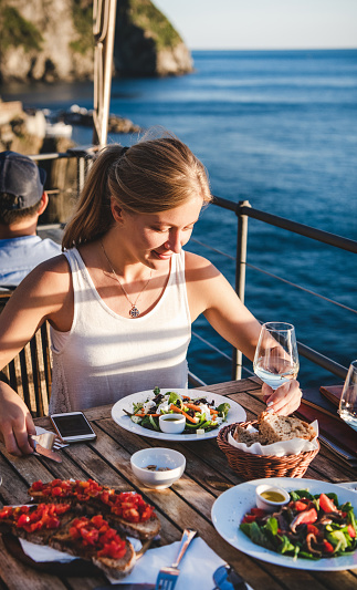 Beautiful young blond woman eating salad and bruschetta and drinking wine in summer open-air cafe in Italy with picturesque sea view background. Travel, wanderlust, Italian lifestyle concept
