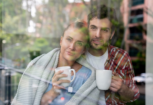 Portrait of a young couple at home drinking a cup of coffee and looking through the window â lifestyle concepts