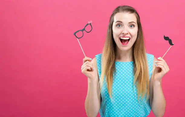 Young woman holding paper glasses and mustache party sticks on a pink background