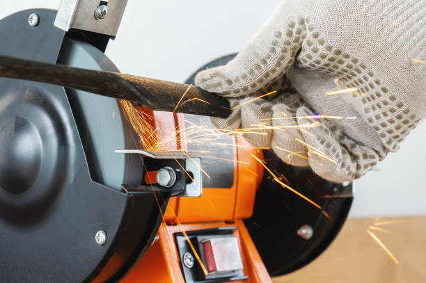 afilar la hoja de un cortacésped con un afilador eléctrico. las manos en los guantes de trabajo sujetan y afilan la cuchilla - afilado fotografías e imágenes de stock