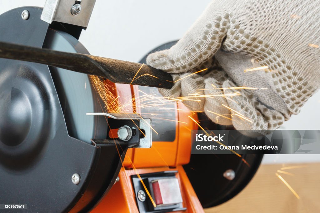 Sharpening the blade of a lawn mower with an electric sharpener. Hands in work gloves hold and sharpen the blade Sharpening the blade of a lawn mower with an electric sharpener. Hands in work gloves hold and sharpen the blade. Sharpening Stock Photo