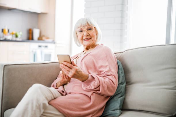 istock Smiling old lady sitting on couch and holding smartphone 1209678296
