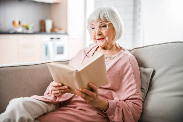 senior woman in glasses reading book at home - woman with glasses reading a book imagens e fotografias de stock