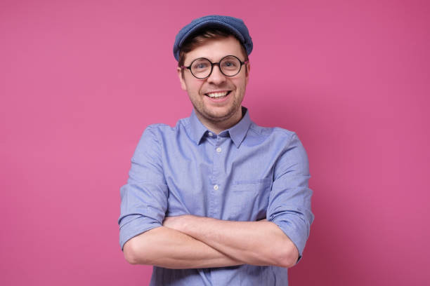 istock Happy caucasian man in glasses and hat with folded hands on pink background. 1209685806