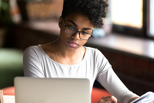 Thoughtful millennial african American woman in glasses look at laptop screen pondering considering making notes, pensive biracial young female student thinking studying online writing using computer