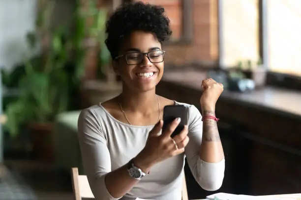 Excited african american millennial tattoo girl in glasses hold smartphone feel euphoric reading good news, overjoyed happy black biracial young woman use cellphone triumph win lottery online
