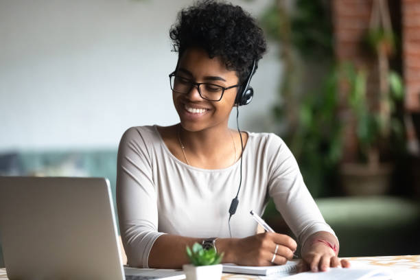 Smiling biracial female in earphones studying making notes Smiling african American millennial female student in headphones and glasses sit at desk watch webinar making notes, happy biracial young woman in earphones work study using computer write in notebook translation photos stock pictures, royalty-free photos & images