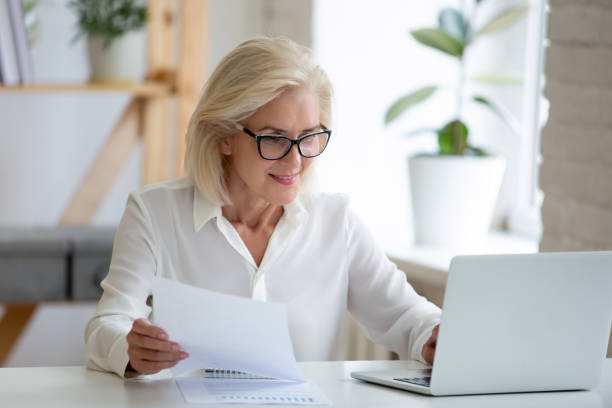 mujer de negocios de mediana edad escribiendo en el pc sostiene papel prepara el informe - office supply desk business person human age fotografías e imágenes de stock