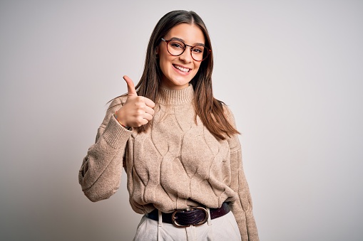 Young beautiful brunette businesswoman wearing casual sweater and glasses standing doing happy thumbs up gesture with hand. Approving expression looking at the camera showing success.