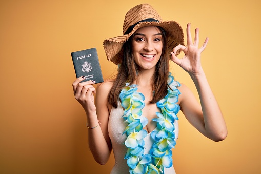 Young beautiful tourist woman on vacation wearing swimsuit and hawaiian lei holding passport doing ok sign with fingers, excellent symbol