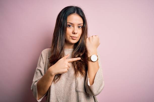 giovane bella donna bruna che indossa un maglione casual in piedi su sfondo rosa in fretta indicando il tempo di guardare il tempo, impazienza, guardando la fotocamera con espressione rilassata - checking the time women impatient wristwatch foto e immagini stock