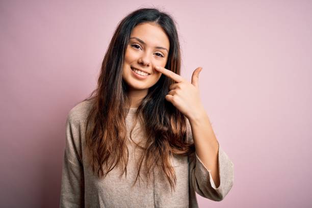 молодая красивая брюнетка женщина носить случайный свитер стоял над розовым фоном указывая пальцем на руку к лицу и носу, улыбаясь веселый. - finger in face стоковые фото и изображения