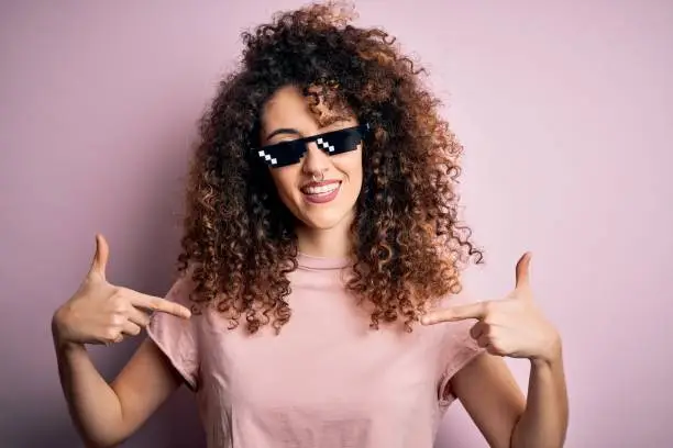 Young beautiful woman with curly hair and piercing wearing funny thug life sunglasses looking confident with smile on face, pointing oneself with fingers proud and happy.