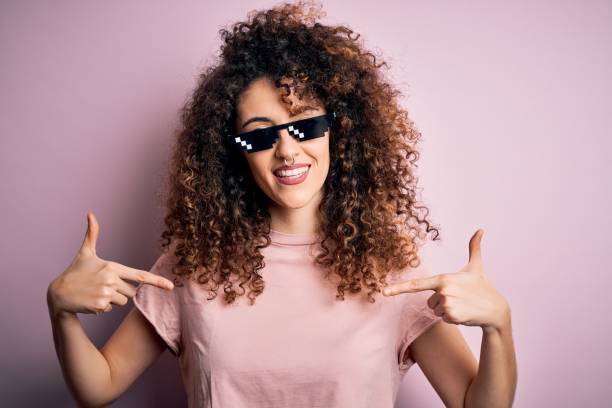 jovem linda mulher com cabelo encaracolado e piercing usando óculos de sol engraçados vida de bandido parecendo confiante com sorriso no rosto, apontando-se com os dedos orgulhosos e felizes. - hooligan - fotografias e filmes do acervo