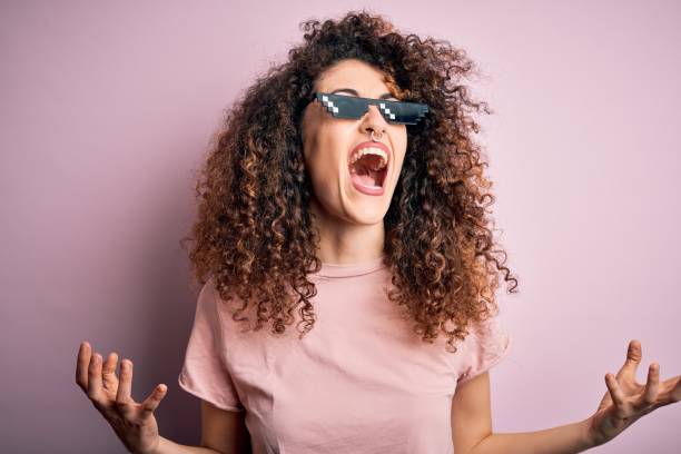 joven mujer hermosa con el pelo rizado y piercing usando divertidas gafas de sol de la vida matón locos y locos gritando y gritando con expresión agresiva y los brazos levantados. concepto de frustración. - bizarre women portrait pierced fotografías e imágenes de stock