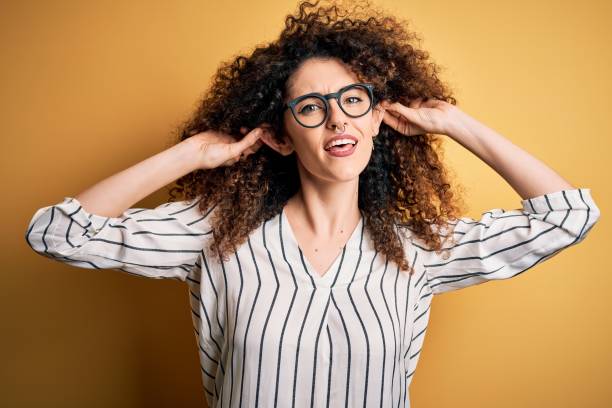 jeune belle femme avec les cheveux bouclés et le piercing portant la chemise rayée et les lunettes souriant tirant des oreilles avec des doigts, geste drôle. problème d’audition - casting glass photos et images de collection