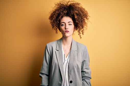 Young beautiful businesswoman with curly hair and piercing wearing elegant jacket with serious expression on face. Simple and natural looking at the camera.