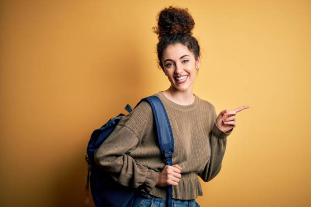 jeune belle femme d’étudiant avec les cheveux bouclés et le sac à dos de port de piercing très heureux pointant avec la main et le doigt au côté - pierced photos et images de collection