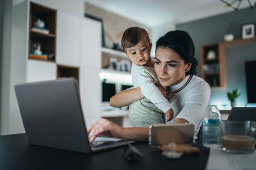 Young modern mother with a baby trying to work on laptop at home and looking tired
