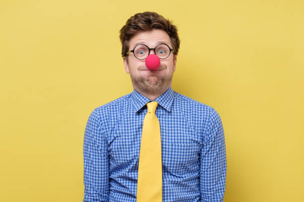 Young handsome man with red clown nose blowing cheeks. Young handsome man with red clown nose blowing cheeks. Studio shot on yellow wall clowns nose stock pictures, royalty-free photos & images