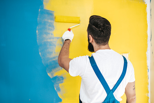 Young handsome bearded man renovating apartment interior. He painting a wall in blue or teal and yellow colors.