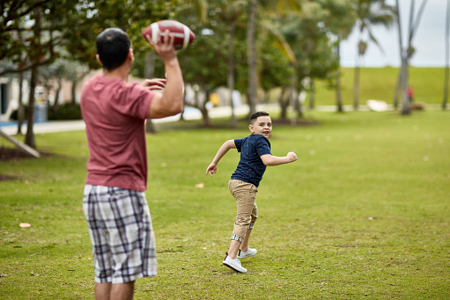 Focus on running young Hispanic American boy looking back over his shoulder as father prepares to pass football in South Florida public park.