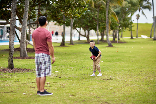 Focus on 6 year old Hispanic American boy facing father after catching football while enjoying healthy weekend exercise in South Florida public park.